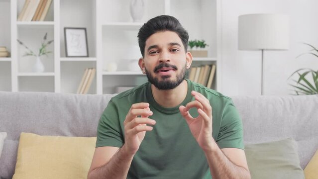 Pleasant bearded man in casual attire sitting on grey couch and talking during video call. Attractive indian male making active hands gesture while having online conference.