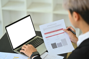 Cropped shot of investment analyst holding saving goal document and using laptop at desk