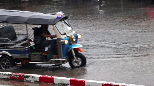 A traditional motorized tricycle - tuk tuk drives through a puddle on the road in rain, Thailand