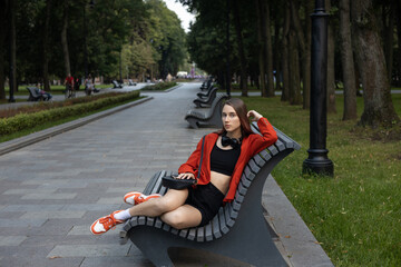 Beautiful girl in stylish red jacket and black suit with headphones sits on park bench and looking at the camera. Generation Z. Relaxation, romantic mood. Student resting after her studies