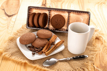 Chocolate cookies, cinnamon, star anise on ceramic saucer with cup and jute napkin on wooden table.