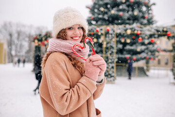 A beautiful curly girl with Christmas candies in her hands poses against the background of a New Year tree on a beautiful snowy winter day.