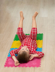 Sport and health concept. A little boy 4 years old in red checkered pajamas is working out on a multi-colored massage orthopedic mat with spikes in a home interior. Close-up.