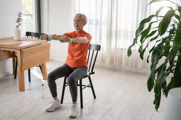 Smiling beautiful senior woman health instructor doing chair exercises with dumbbells - obrazy, fototapety, plakaty