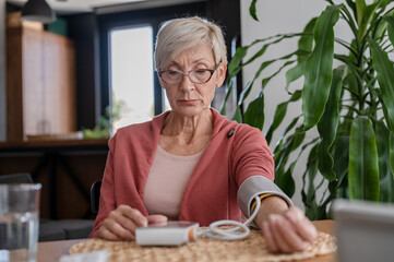 Older woman sitting alone at home, measuring her blood pressure with a home device - obrazy, fototapety, plakaty