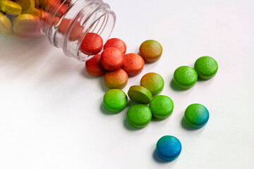 Colored round candies, yellow, red, green and blue, spilling from a glass vial, on a white background. They roll out of the pot