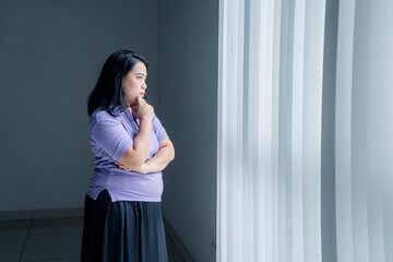 Image of obese woman looks pensive while standing by the window