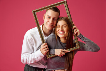 Portrait of happy young couple, man and woman, posing with picture frame against pink studio...