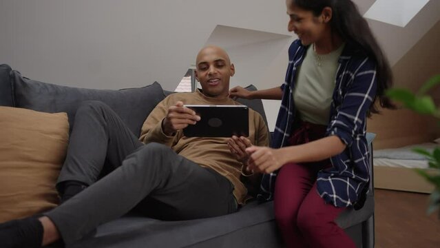 Husband lying on the couch invites his wife to come and see something on his tablet 