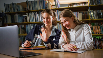 Two girl students sitting in the library and using a laptop during virtual online lessons. Internet, education, and e-learning concept.