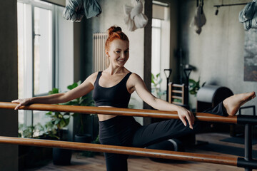 Fototapeta premium Happy young woman good stretch posing in fitness studio during barre workout