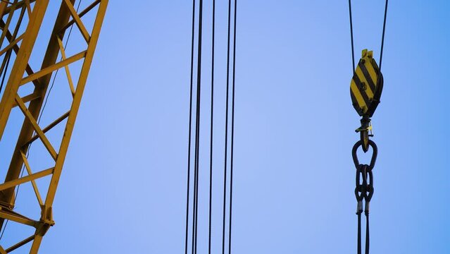 Steel cables and hooks are hanging from sturdy boom of crane at arbitrary unspecified location against blue sky. Yellow construction crane for cargo loading