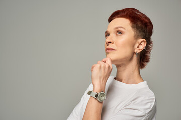 thoughtful redhead non-binary person holding hand near chin and looking away on grey backdrop