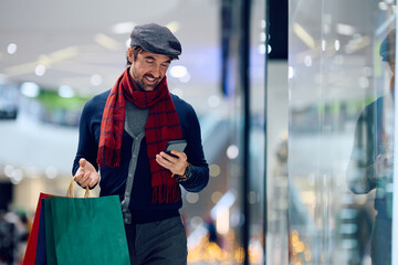 Happy man using cell phone while walking with shopping bags at mall.