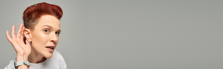 curious redhead bigender person holding hand near ear while looking at camera on grey, banner