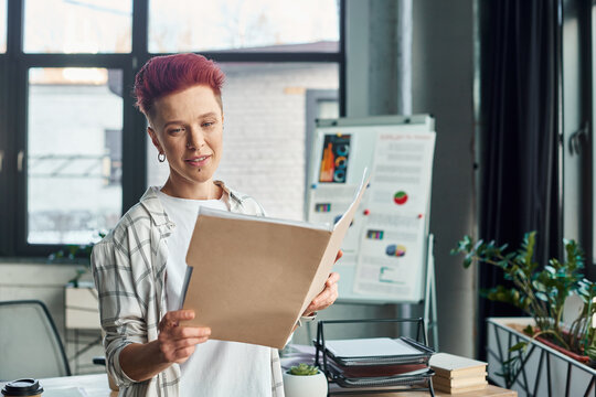 stylish bigender person in casual attire looking in folder with documents in contemporary office - Powered by Adobe