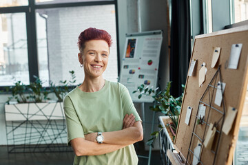 happy non-binary person with short hair looking at camera near corkboard with paper notes in office