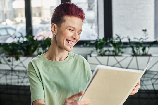 joyful queer person with short hair looking  at folder with documents in contemporary office