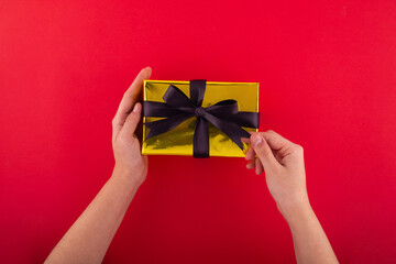 First person top view photo of hands unpacking gold paper gift box with black satin ribbon bow over on isolated red background with blank space.