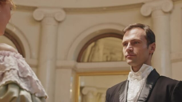 Waist up slowmo of handsome young Caucasian gentleman in elegant tuxedo suit approaching to young lady and asking her to dance with him at ball
