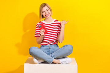 Full size portrait of lovely positive girl sit podium cube hold smart phone direct finger empty space isolated on yellow color background