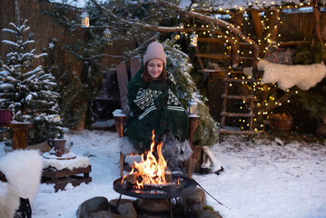 Young attractive woman in winter clothes sitting in a armchair, warming herself by the fire and relaxing outside in winter