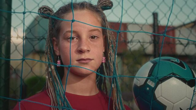 Confident adolescent girl emphasizes passion for football posing with ball on soccer field. Girl goalkeeper with dyed braids enjoys playing football after school at local playground for football