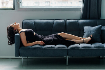 Young beautiful brunette girl in a black dress sitting on the sofa - obrazy, fototapety, plakaty