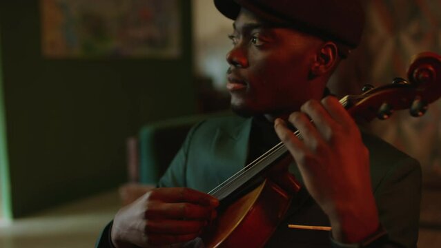 Chest up of young Black male jazz musician in beret hat and emerald suit playing violin before performance in fancy club with dimmed lights
