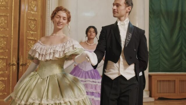Medium slowmo of three young diverse dancing couples looking at each other and holding hands in gloves while walking in row one by one along spacious Victorian style ballroom