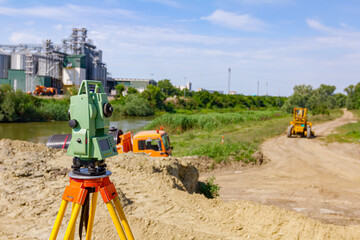 Geodesist device on tripod at building site over water are big grain silos in industrial landscape © Roman_23203