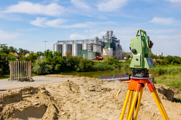 Geodesist device on tripod at building site over water are big grain silos in industrial landscape
