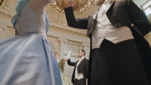Low angle slowmo of two beautiful aristocratic ladies wearing poof ruffled dressed dancing with male partners at debutante ball in luxurious ballroom