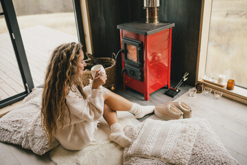 Young woman sitting by the fireplace in white sweater, drinking tea in cozy log cabin. - obrazy, fototapety, plakaty