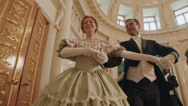 Dollying slowmo shot of young diverse members of aristocracy wearing medieval dresses and suits dancing in pairs at pompous ball party indoors