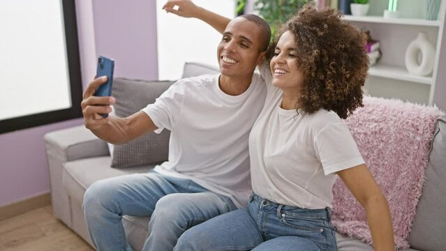 Beautiful couple enjoying fun moments together at home, confidently taking joyful selfie picture on sofa using smartphone.