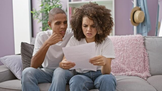 Worried beautiful couple sit on sofa, reading problematic document at home, stressed expressions painted over their faces