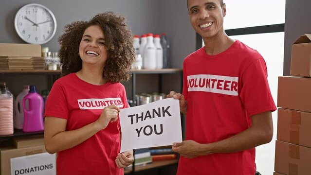 Young man and woman volunteers, smiling with gratitude, holding a 'thank you' message banner at the charity center amidst donations