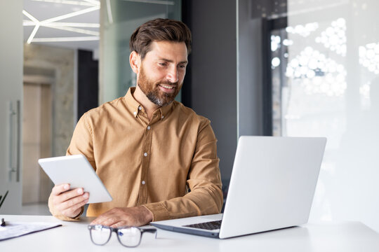 A young male programmer and developer sits in the office at the table and works on a laptop and with a tablet. Talks on a video call - Powered by Adobe
