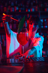 Young beautiful woman, bartender in modern bar, making delicious Aperol Spritz cocktail in neon, decorating with leaf. Concept of occupation, nightlife, bar, party, alcohol drink, mixologist
