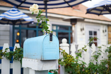 A classic designed blue mailbox in front of the house fence and flower garden (as blurred background). Close-up and selective focus.