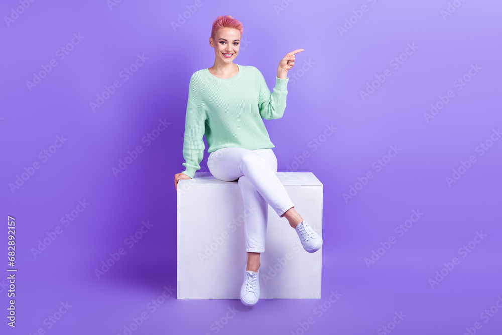 Wall mural Full length photo of cute cheerful lady dressed teal pullover sitting white platform pointing empty space isolated purple color background