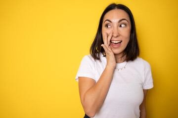 A close up portrait of a young pretty woman telling secret information, holding her hand near mouth and standing against yellow background - obrazy, fototapety, plakaty