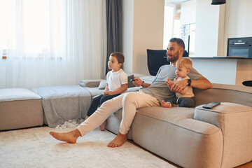 Father with two baby boys is sitting at sofa