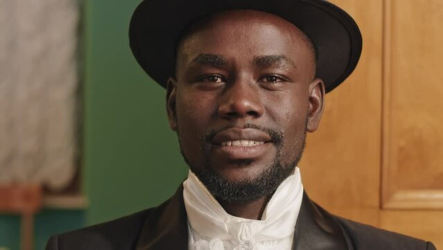 Tilting up portrait of aristocratic young Black man wearing black tuxedo suit and cylinder hat posing for camera in classic ball hall