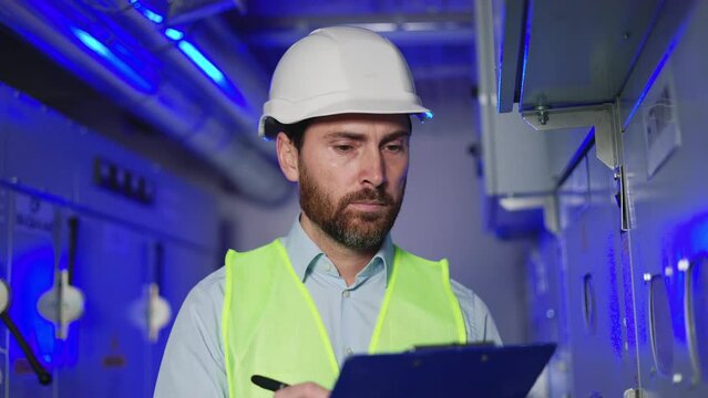 Professional engineer in protective clothing taking notes on clipboard at electrical substation Man checking data and meter readings in technical room of factory. Distribution electrical switchboards.