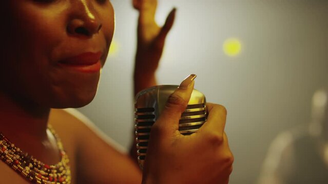 Medium closeup of young African American female jazz singer holding retro microphone and singing while giving concert in fancy jazz club in evening