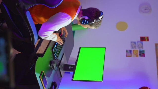 vertical shot, girl with short hair puts on her headphones to play a video game on her computer. Green screen for adaptations. High quality 4k footage