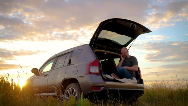 Tired lazy napping man male manager worker bored at work project online in notebook computer sitting in car trunk while traveling, general plan in field at sunset. Busy worker freelancer entrepreneur