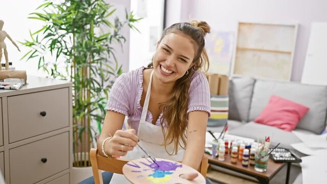 Smiling and confident, beautiful young hispanic woman artist skillfully holding her paintbrush and palette in bustling art studio setting